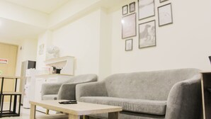 A living room with a grey sofa, a wooden coffee table, and a small table with a remote control. There are framed pictures on the wall and a kitchen area in the background.
