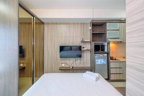 A modern hotel room with a large bed, a flat-screen TV mounted on the wall, a small kitchen area with a refrigerator and microwave, and a sliding door closet.