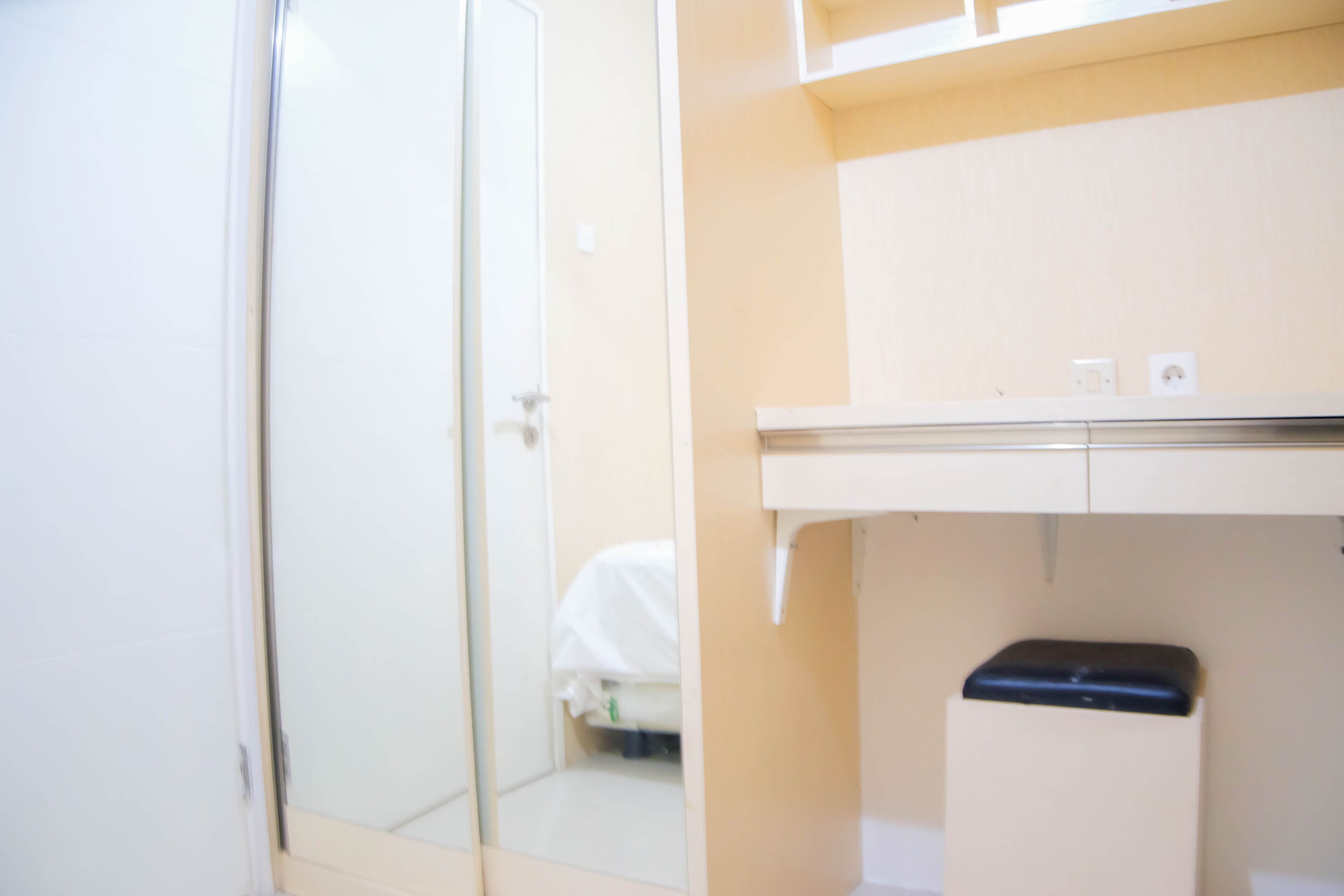A room with a closed door, a bed with a white cover, a small trash bin, and a light switch on the wall.