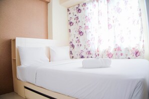A neatly made bed with white linens and a folded towel, a floral curtain, and a plain wall.