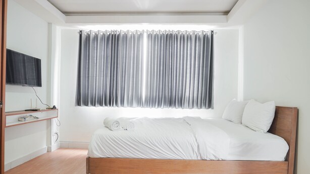 A bedroom with a wooden bed, white bedding, a television on a shelf, and a window with grey curtains. - Scenic And Strategic Studio Amethyst Apartment (Jakarta)