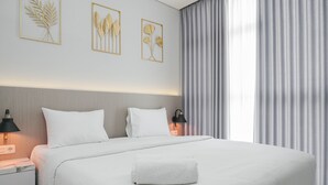 Een net opgemaakt bed met witte lakens, een wit kussen en een opgerolde witte handdoek. Er staat een nachtkastje met een lamp en een stopcontact. De kamer heeft een grijze gordijn en een muur met drie kunstwerken in lijsten.