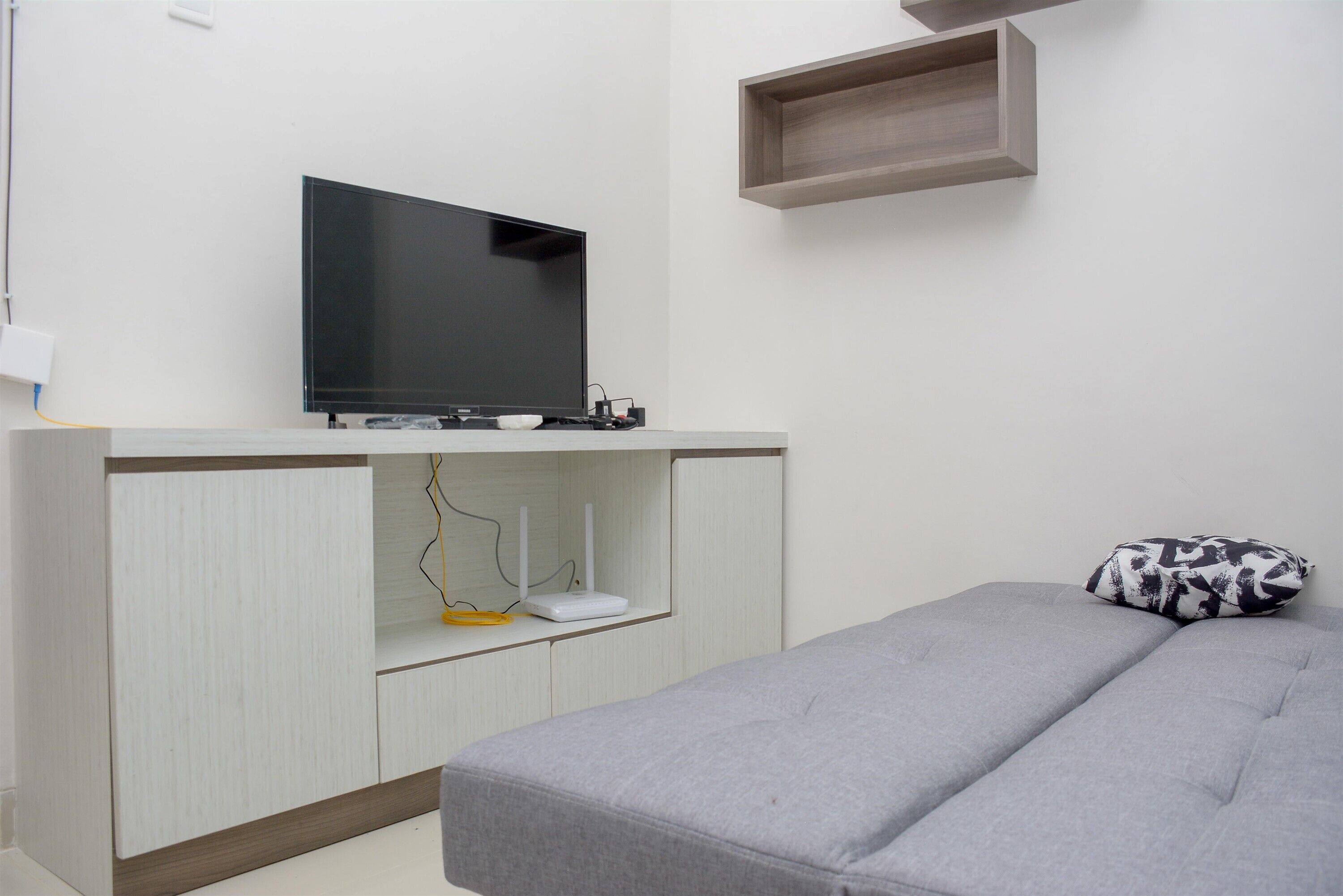Une pièce moderne avec un canapé gris, un téléviseur à écran plat posé sur un meuble blanc et une petite étagère en bois.