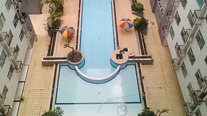 A rectangular swimming pool surrounded by a tiled walkway, with multiple umbrellas and potted plants. - Spacious Studio Apartment At Jarrdin Cihampelas (Bandung)