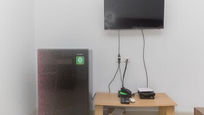 A room with a flat-screen TV mounted on the wall, a refrigerator, and a wooden table with a projector and remote controls. - Homey And Comfort Studio At The Enviro Apartment (Cikarang)