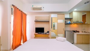 A hotel room with a bed, a TV, a desk with a computer, a kitchen area, and an air conditioning unit. - Homey And Simply Studio Room At Margonda Residence 3 Apartment (Depok)