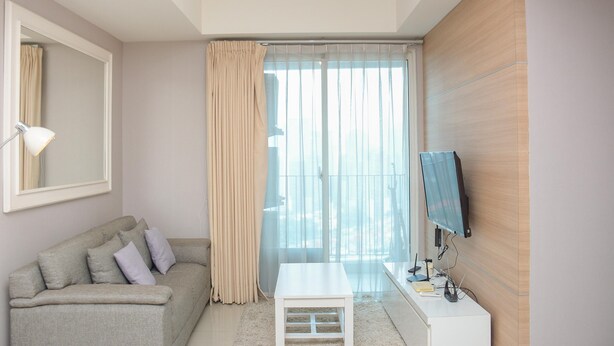 A modern hotel room with a grey sofa, a small white table, a TV on a wooden wall, and a large window with sheer curtains.
