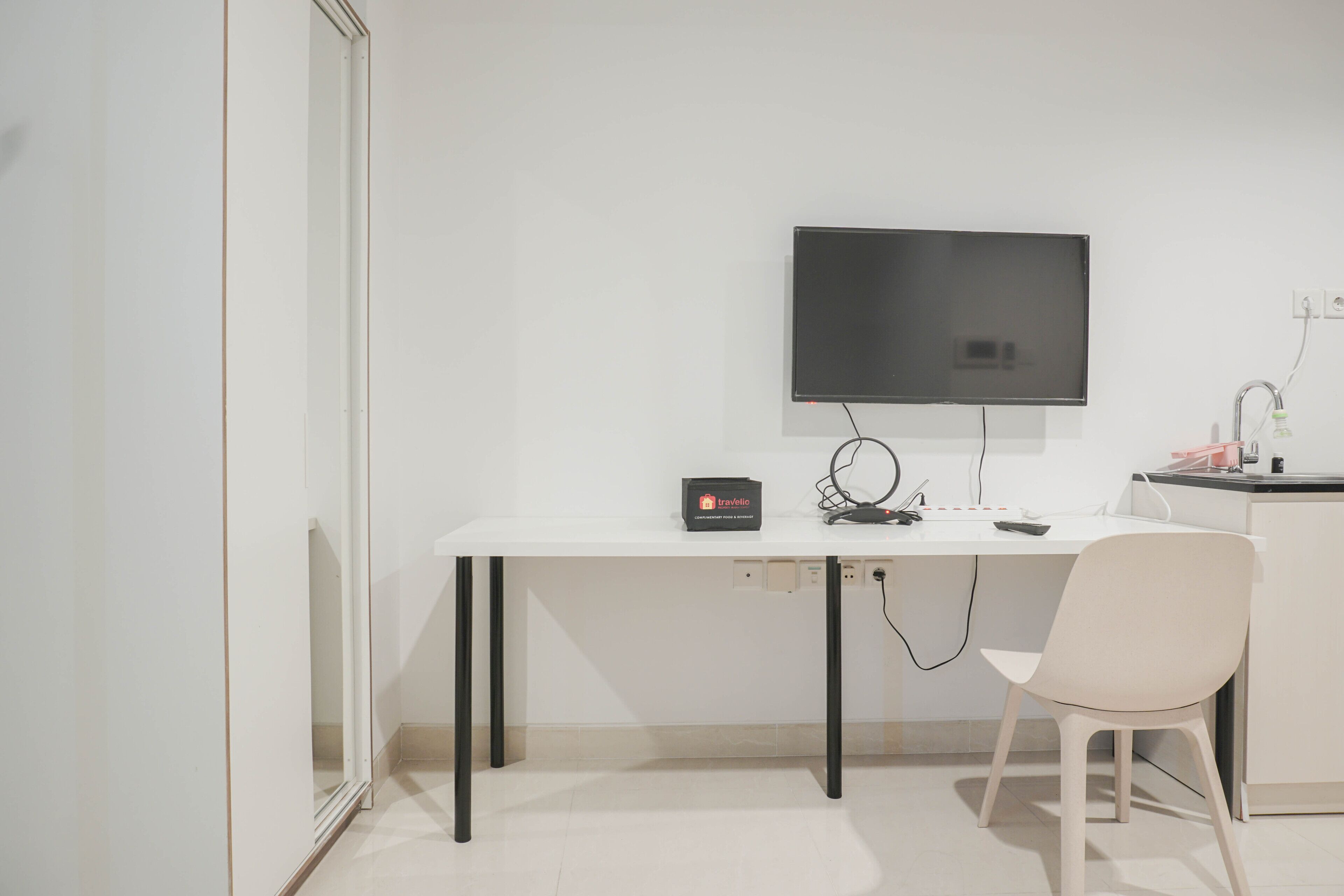 A modern, minimalist office space with a white desk, a chair, a flat-screen TV mounted on the wall, and a small travel-related device on the desk.
