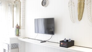 A modern living room with a mounted flat-screen TV, a small shelf with a box labeled "Travelio," and decorative wall art. - Elegant & Stylish Studio At Gateway Pasteur Apartment (Cimahi)