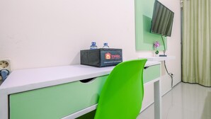 Quarto de hotel com escrivaninha branca, gavetas verdes, uma cadeira verde, televisão de tela plana presa à parede e uma janela com cortinas verdes.