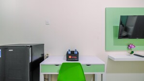 Um quarto pequeno com uma mesa, uma cadeira, um refrigerador, uma prateleira com controle remoto e uma televisão.