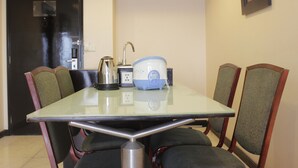 A dining area with a glass table, four chairs, a kettle, a coffee maker, and a power outlet.