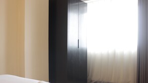 A hotel room with a bed, a remote control on the bed, a dark wooden wardrobe, and sheer curtains on the window.