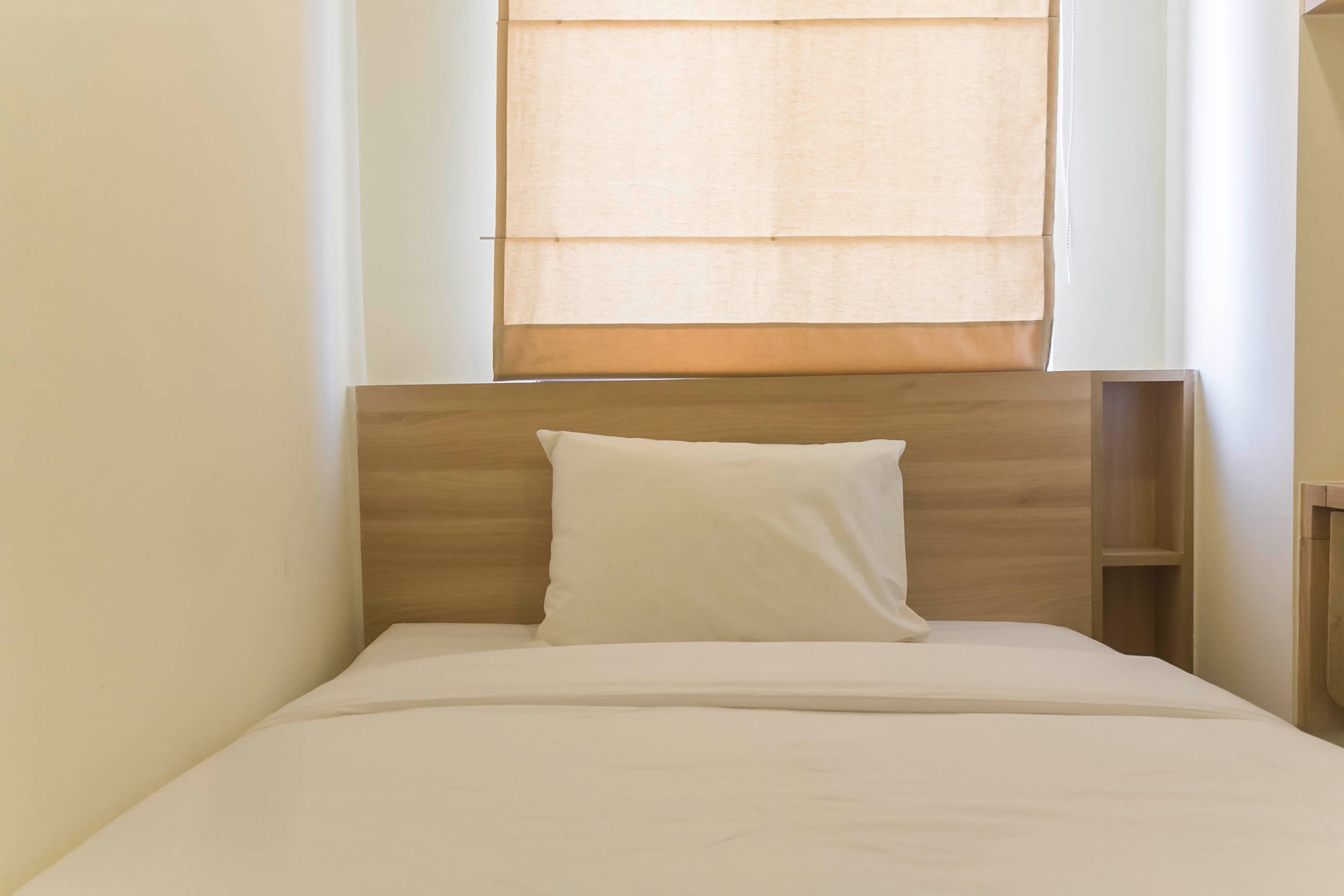 A small, neatly made bed with a wooden headboard and a white pillow, positioned under a window with sheer curtains.