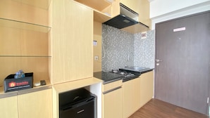 A compact kitchen with a black stove, a microwave, and a small trash bin. There is a shelf with a box labeled 'Travelio' and a water bottle. The kitchen has wooden cabinets and a tiled backsplash. - Relaxing 2Br Apartment At The Jarrdin Cihampelas (Bandung)