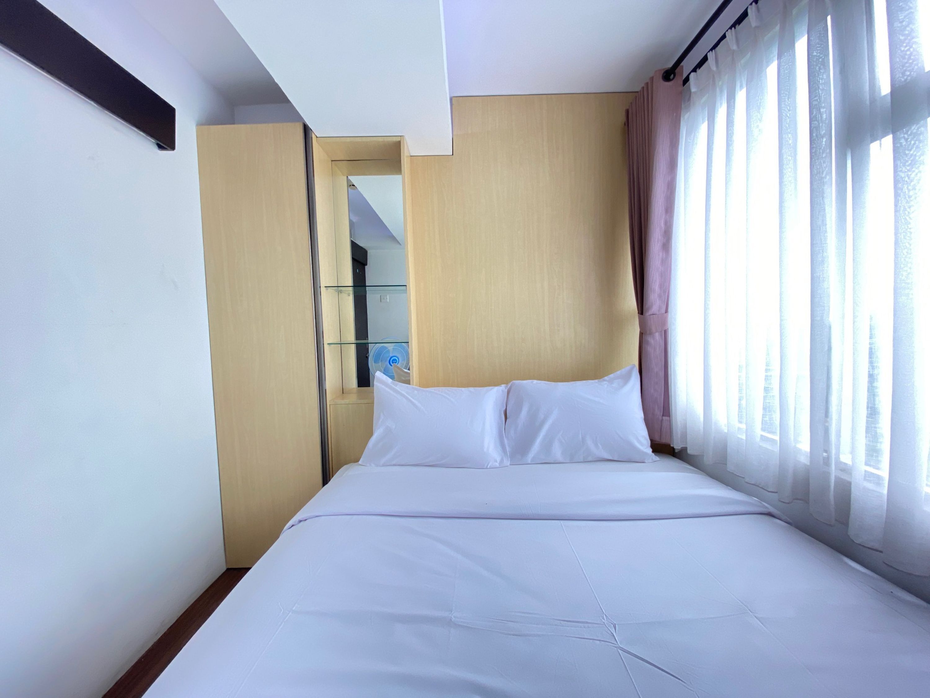 A hotel room with a bed, a wooden wardrobe, a glass shelf, and a window with sheer curtains.