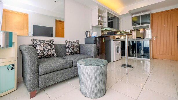 Ein modernes Wohnzimmer mit grauem Sofa, gemusterten Kissen, einem Glastisch und einer Waschmaschine im Hintergrund.