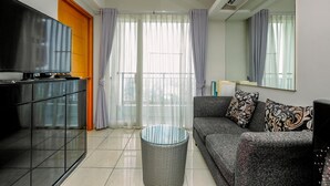 Ein modernes Wohnzimmer mit einem Flachbildfernseher, einem Sofa mit gemusterten Kissen, einem Glastisch und einem großen Fenster mit transparenten Vorhängen.
