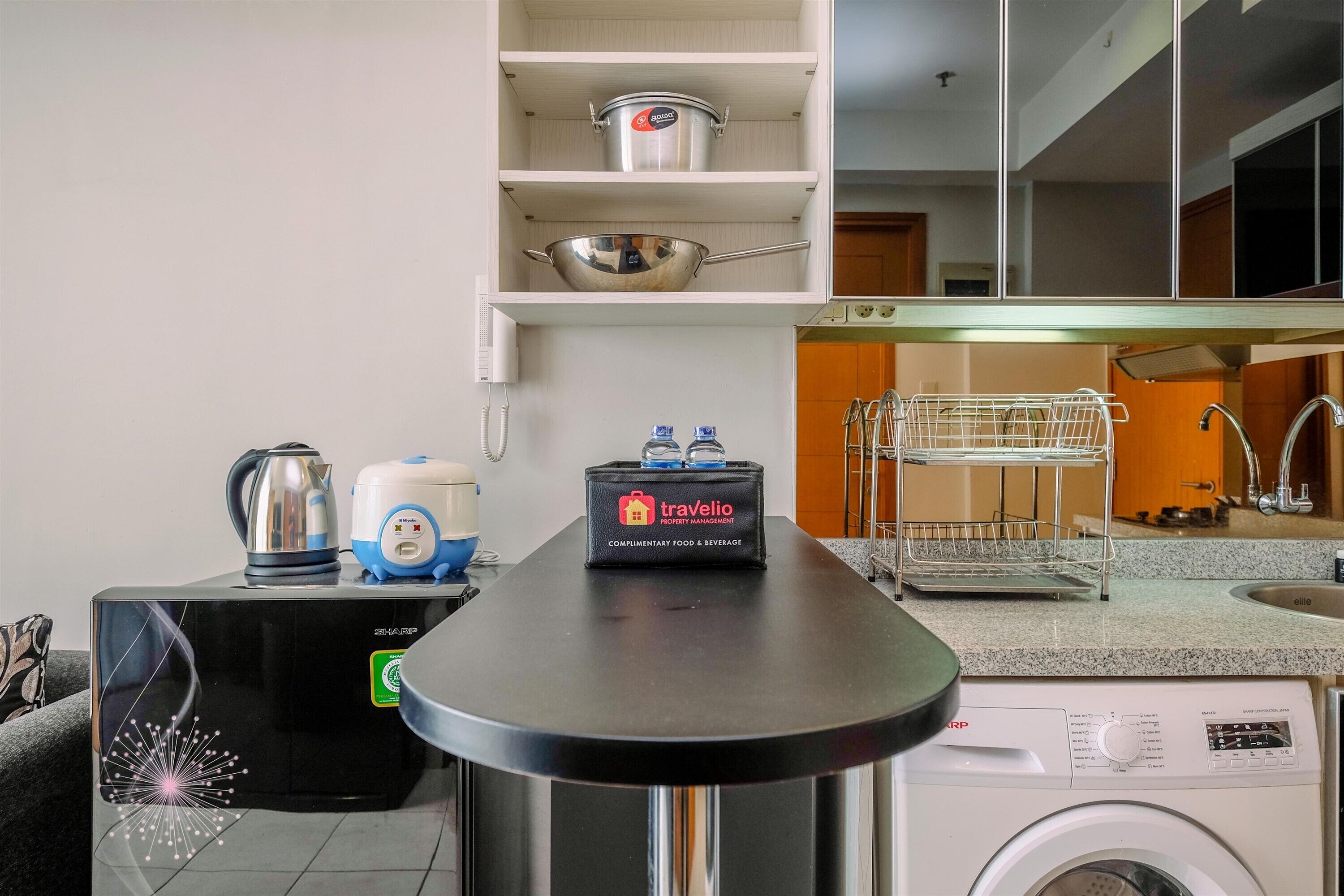A kitchen with a countertop, a washing machine, a kettle, a coffee maker, a travel bag, and a shelf with pots.