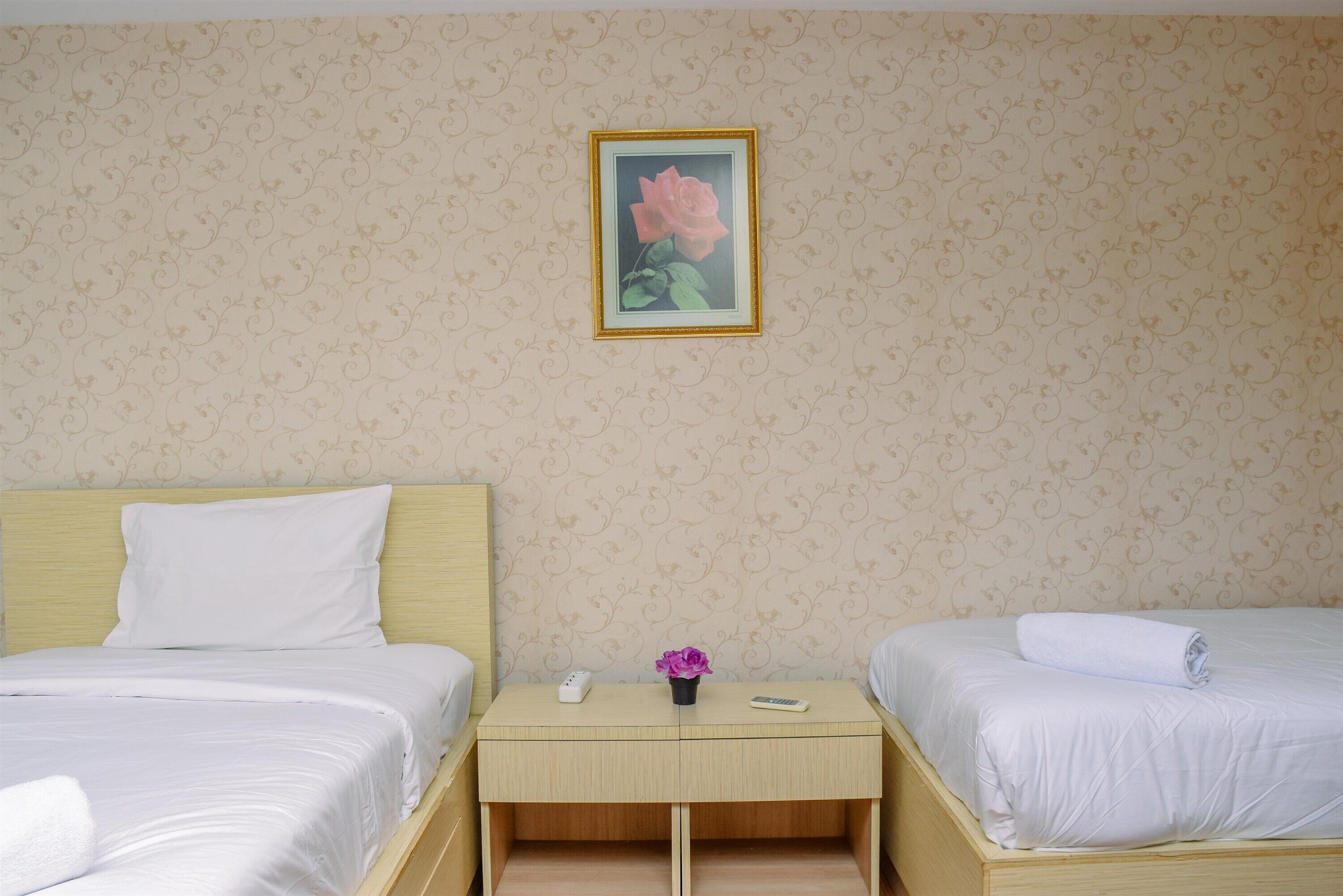 A hotel room with two beds, a bedside table with a remote control, a vase with a flower, and a framed picture on the wall.