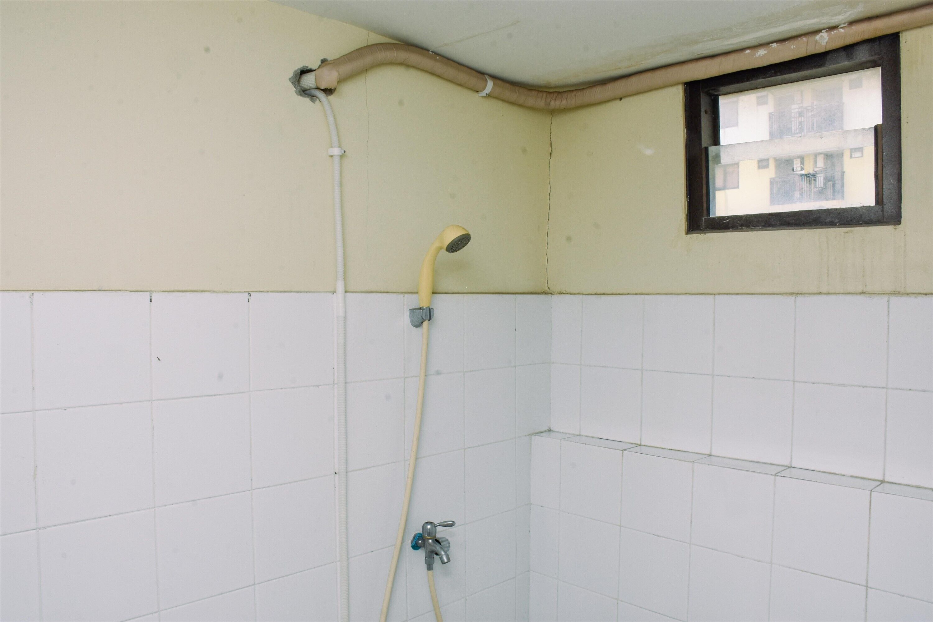 Een badkamer met witte betegelde wanden, een douche met een gele doucheslang en een raam met uitzicht op gebouwen.