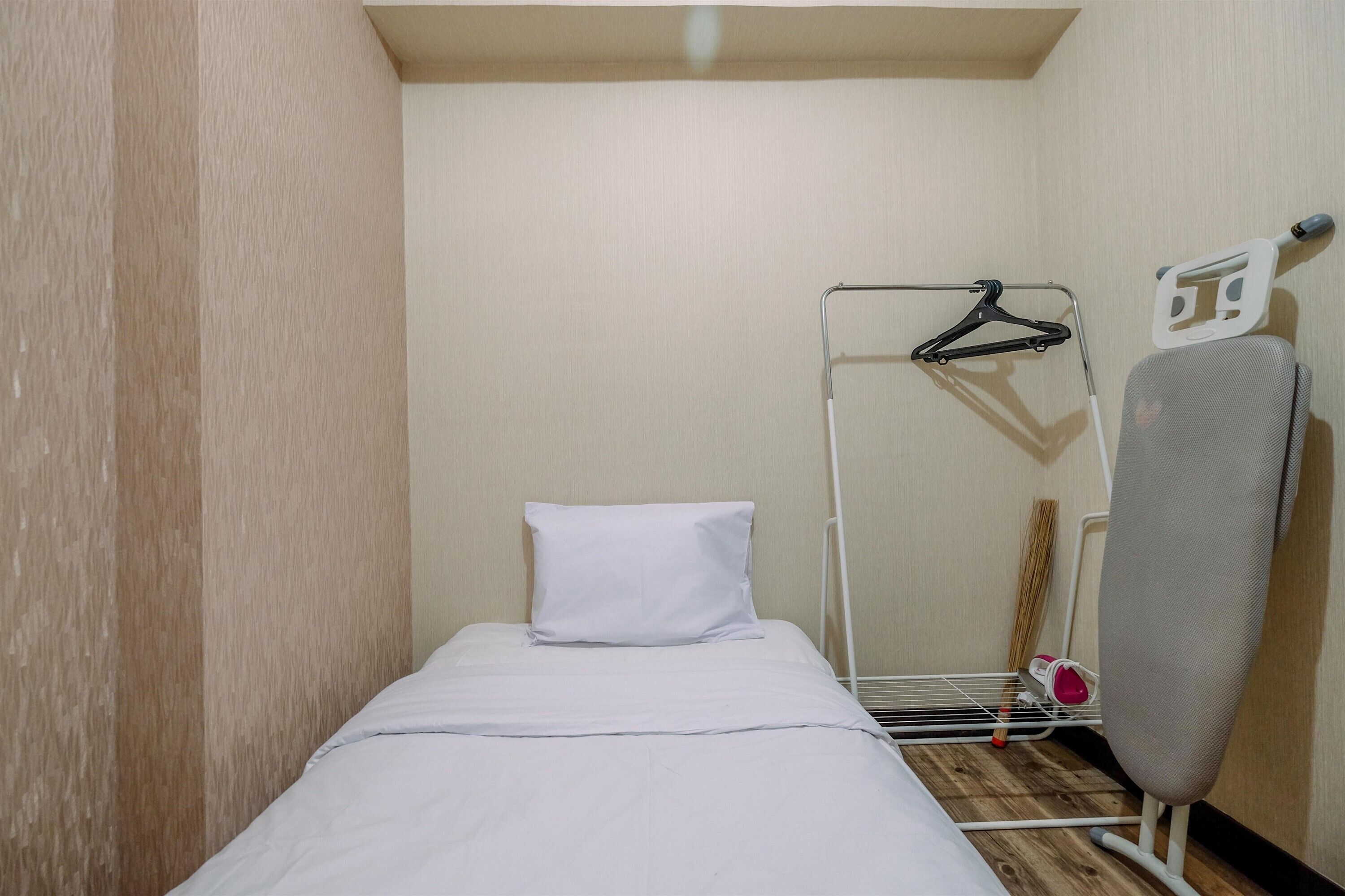 A small, single-bed room with a white bedspread, a white pillow, a metal hanger stand, and a white ironing board.