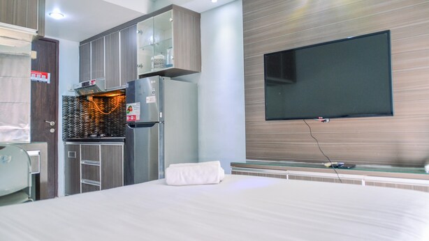 A hotel room with a bed, a TV mounted on the wall, a kitchenette with a stove, and a glass cabinet. - Homey And Simple Studio At Serpong Greenview Apartment (South Tangerang)