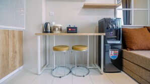 A compact kitchen with a coffee machine, a travel coffee maker, and a refrigerator. There are two stools and a brown sofa in the living area. - Brand New Fabulous 2Br Podomoro Golf View Apartment By Travelio (Gunung Putri)