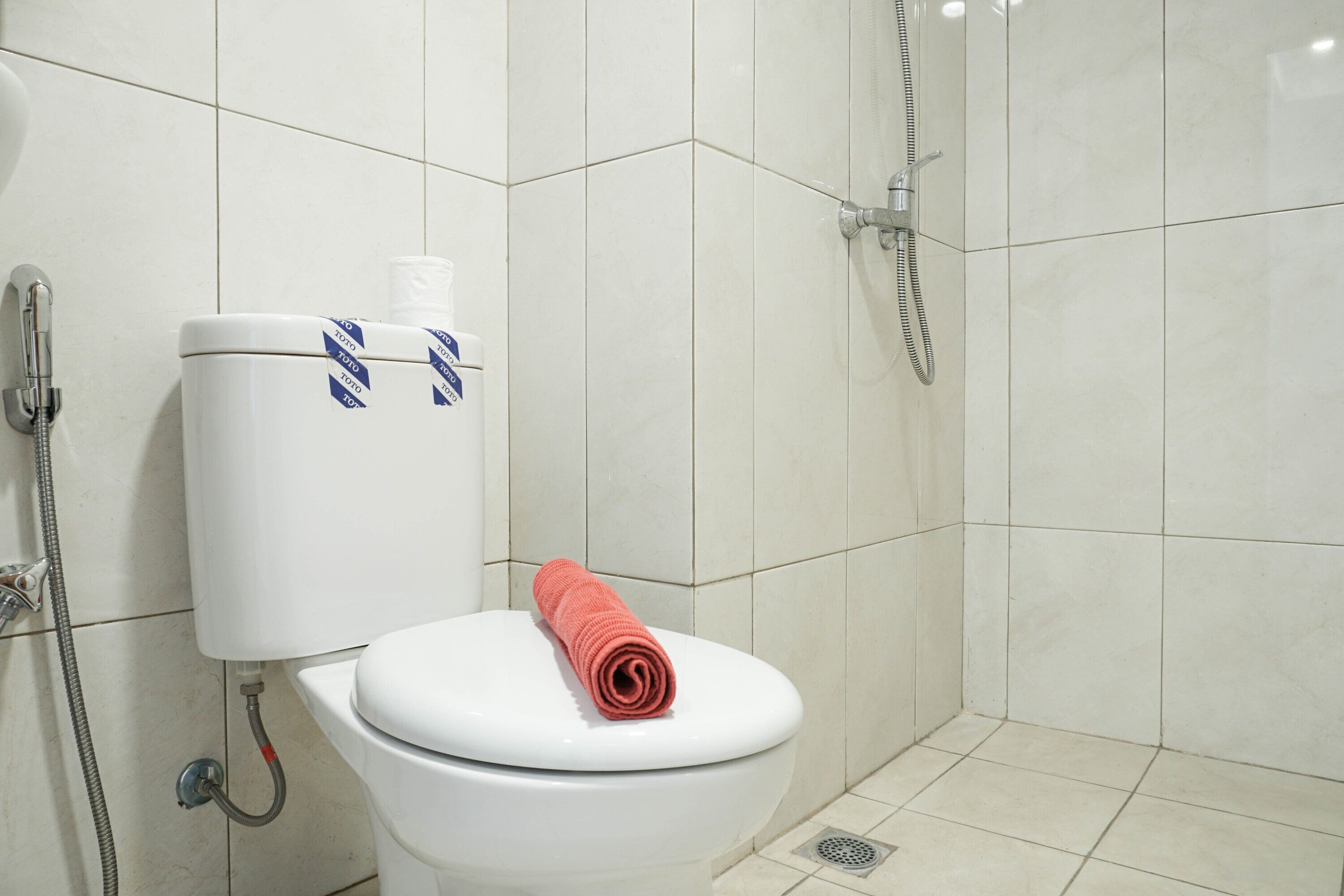 A white toilet with a red towel on the seat, a showerhead, and a toilet paper holder with a blue and white pattern.
