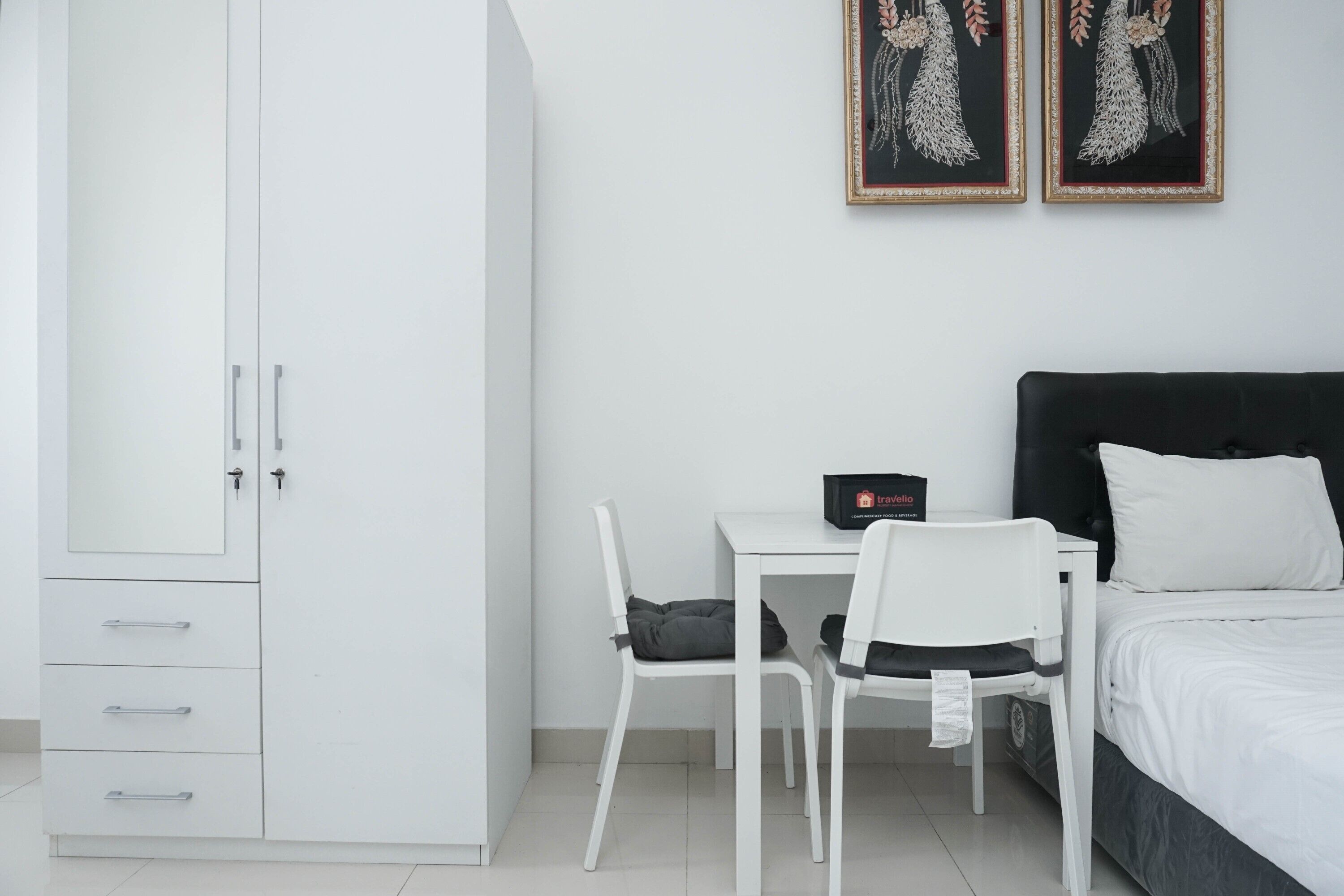 Ein modernes Schlafzimmer mit einem weißen Kleiderschrank, einem Bett mit schwarzem Kopfteil, einem Schreibtisch mit Stuhl und zwei gerahmten Bildern an der Wand.