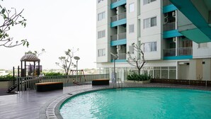 A swimming pool with a wooden deck, a gazebo, and a multi-story building with balconies. - Relax Studio Apartment At The Nest Near Puri By Travelio (Tangerang)