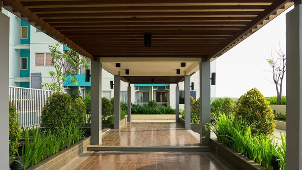 A covered outdoor area with a tiled floor, surrounded by greenery and a building in the background. - Relax Studio Apartment At The Nest Near Puri By Travelio (Tangerang)