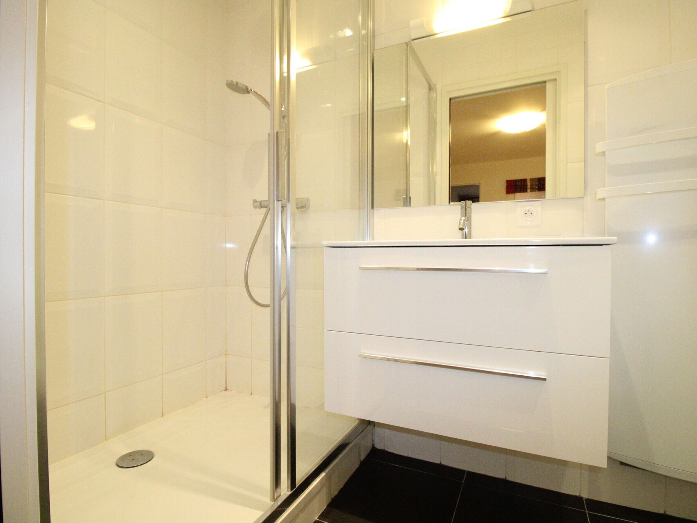 Un baño con cabina de ducha de vidrio, paredes cubiertas de azulejos blancos y un tocador blanco con cajones.