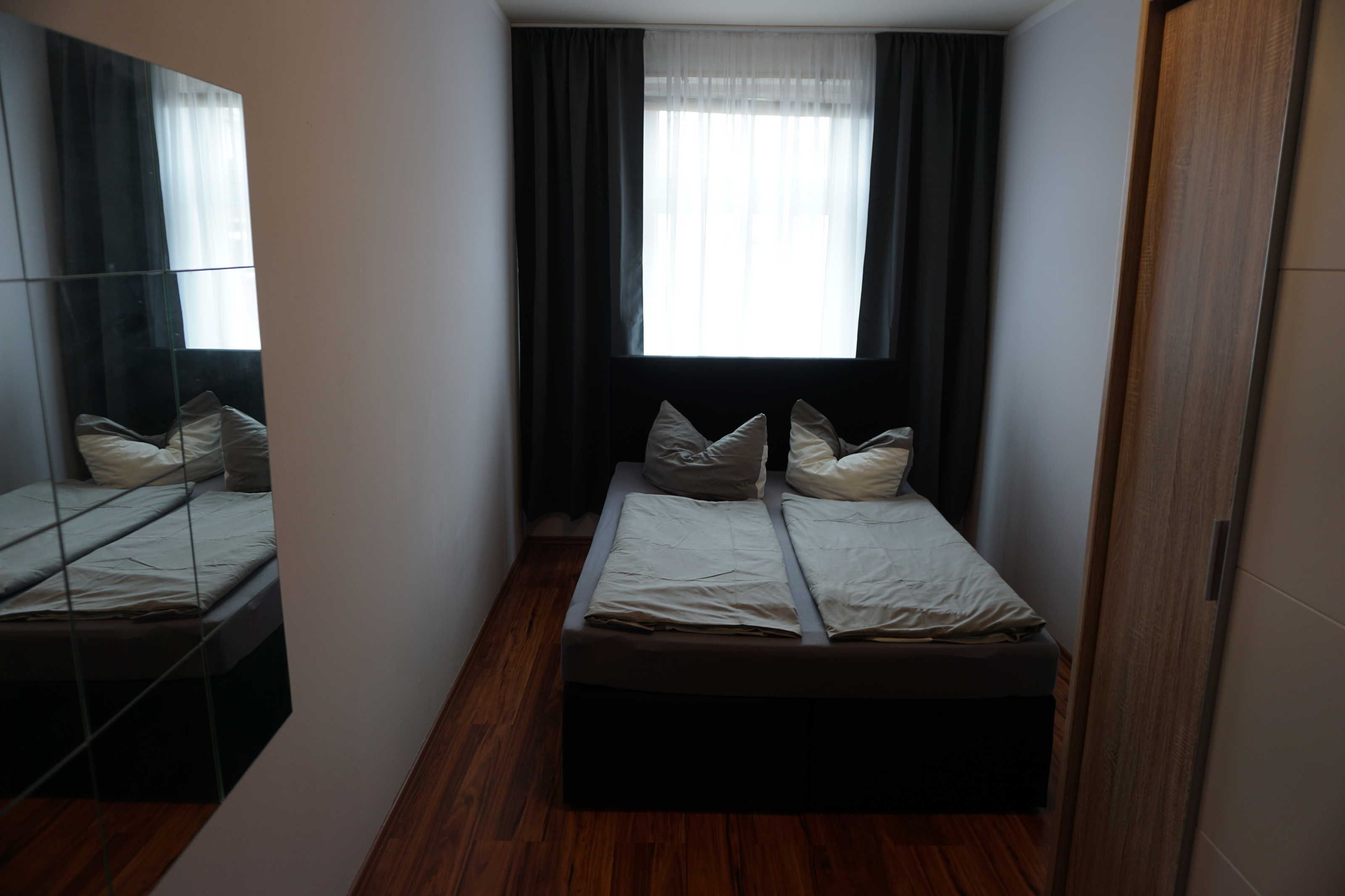 Ein kleiner, schmaler Raum mit einem Bett, einem Spiegel und einem fenster mit Vorhängen.