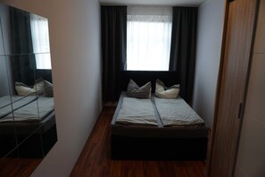 Ein kleiner, schmaler Raum mit einem Bett, einem Spiegel und einem fenster mit Vorhängen.