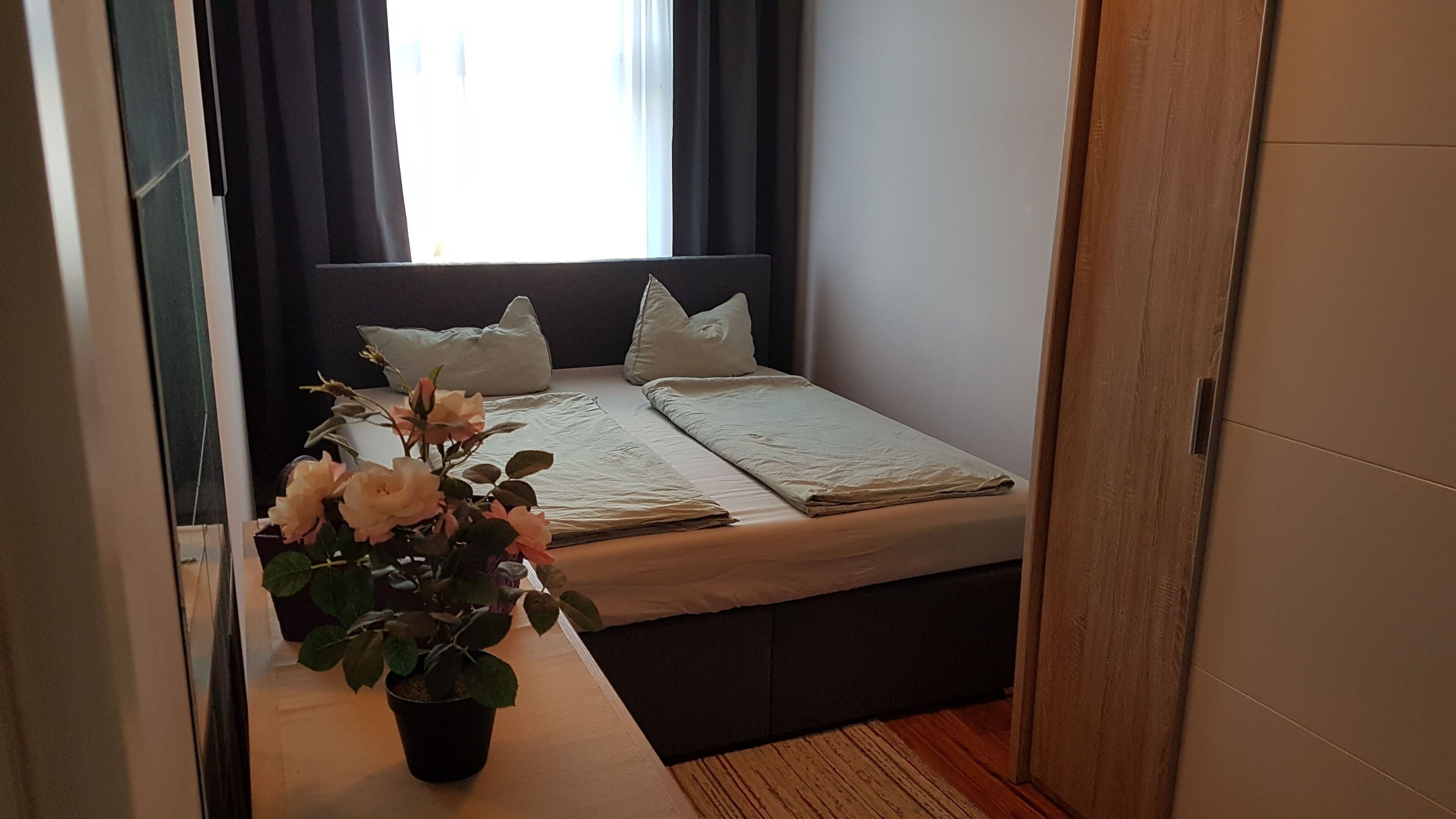 Ein Schlafzimmer mit Bett, Kissen, einem Nachttisch mit Topfpflanze und einem Fenster mit Vorhängen.