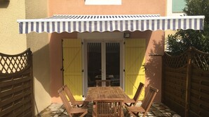 A patio with a wooden table and chairs, a striped awning, and a yellow gate. - Semi-detached house close to the beach (Marseillan Plage)