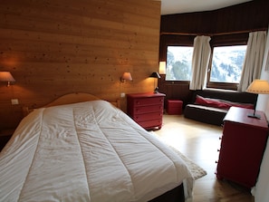 Quarto com painéis de madeira, uma cama, cômoda, mesa de cabeceira e janela com cortinas.