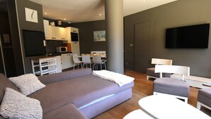 A modern living room with a grey sectional sofa, white armchairs, and a white coffee table. There is a flat-screen TV mounted on the wall, a dining area with a table and chairs, and a kitchen area with a microwave and cabinets. - Avoriaz: Wellness space, 8 pers, 90 sqm (Avoriaz)