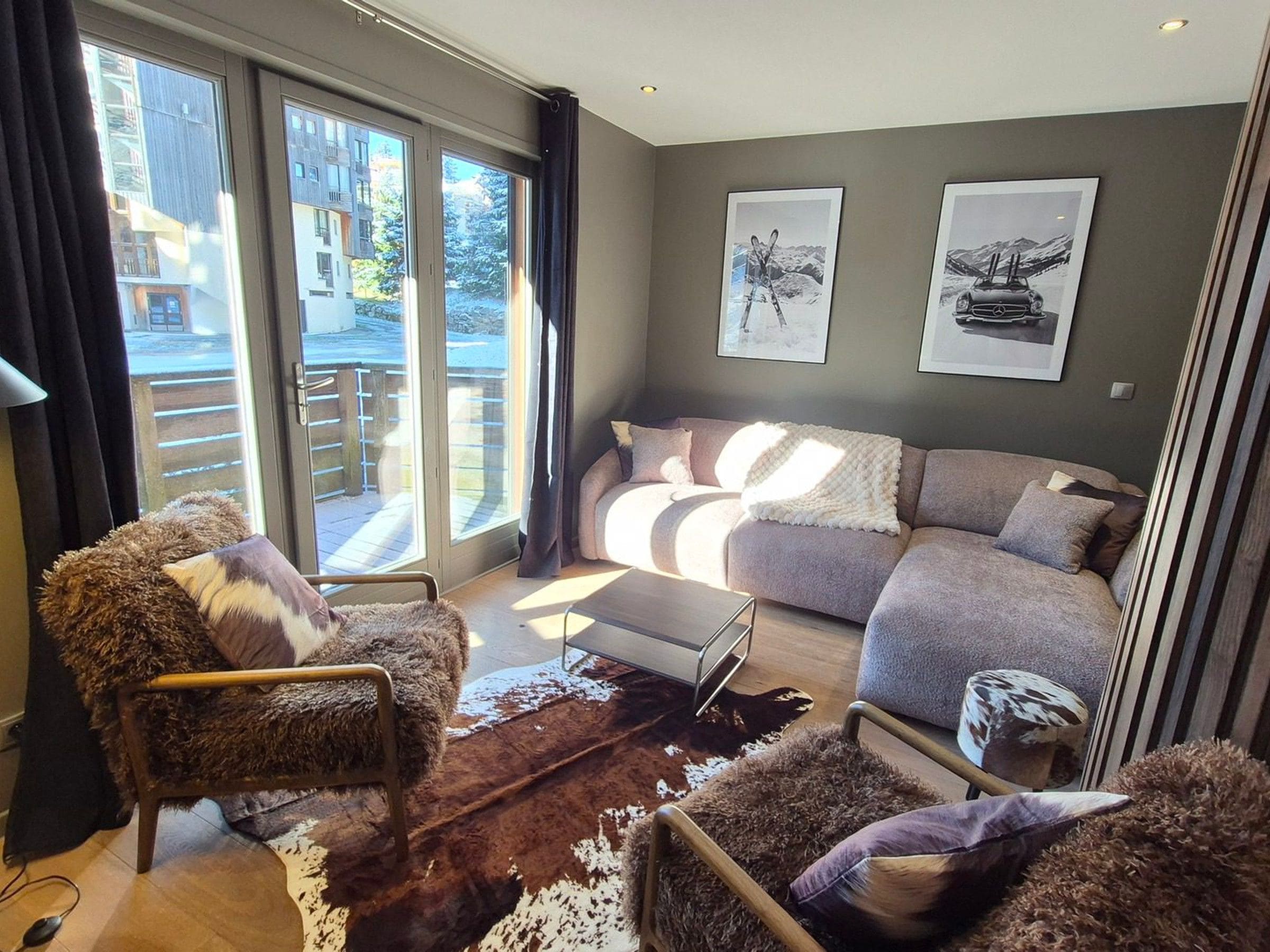 A modern living room with a sofa, armchair, and a coffee table. There are large windows with curtains, and two framed pictures on the wall.