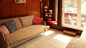A cozy room with a wooden wall, a sofa with patterned cushions, a small table with a lamp, and a window with a view of outdoor seating.