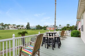A wooden deck with outdoor seating, a white fence, and a view of a green lawn and buildings in the background.