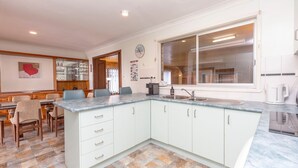 A modern kitchen with a large window, a dining area with chairs, and a kitchen island with drawers. - Wayila - Riverfront - Pet Friendly (Hawks Nest)