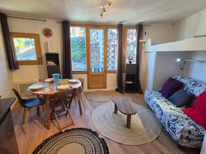 Un salon meublé comprenant un coin repas, un canapé, un téléviseur et une vue sur les immeubles par les fenêtres.