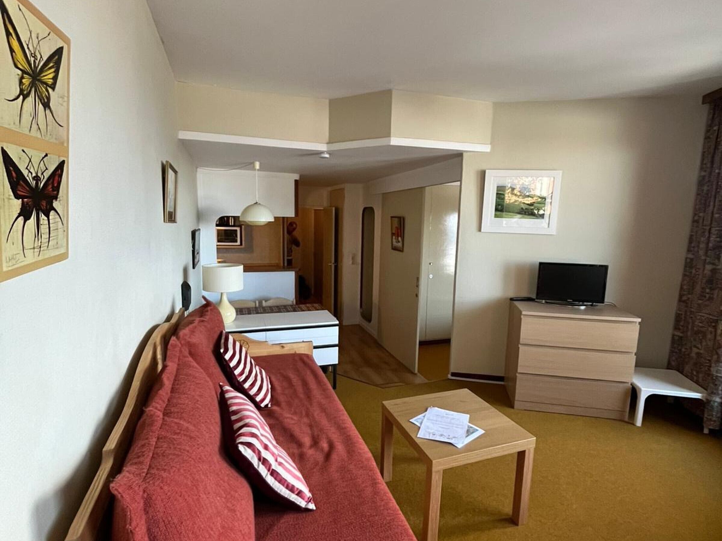 Un espacio habitable compacto con un sofá, una mesita, un televisor y un escritorio. Se ve una cama al fondo y una puerta que da a otra habitación.