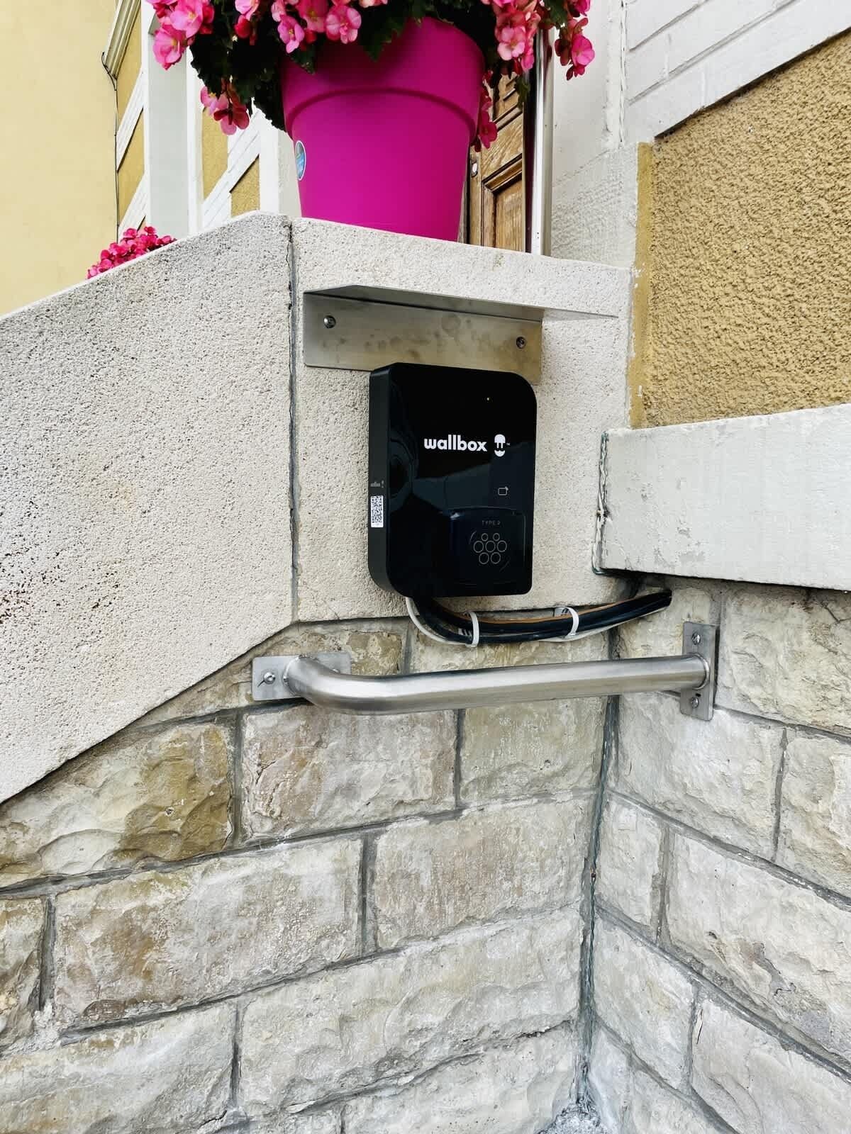 Une borne de recharge murale fixée au mur, dotée d’un code QR, d’une main courante en métal et d’une plante en pot aux fleurs roses.