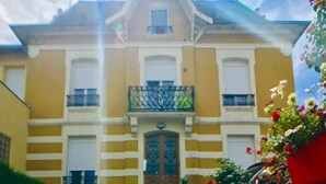 A building with a yellow facade, a central entrance with a double door, and a small balcony with a metal railing. - Romy Schneider (Vittel)