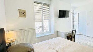 A bedroom with a bed, a desk, a chair, a TV, and a window with blinds. - Romy Schneider (Vittel)