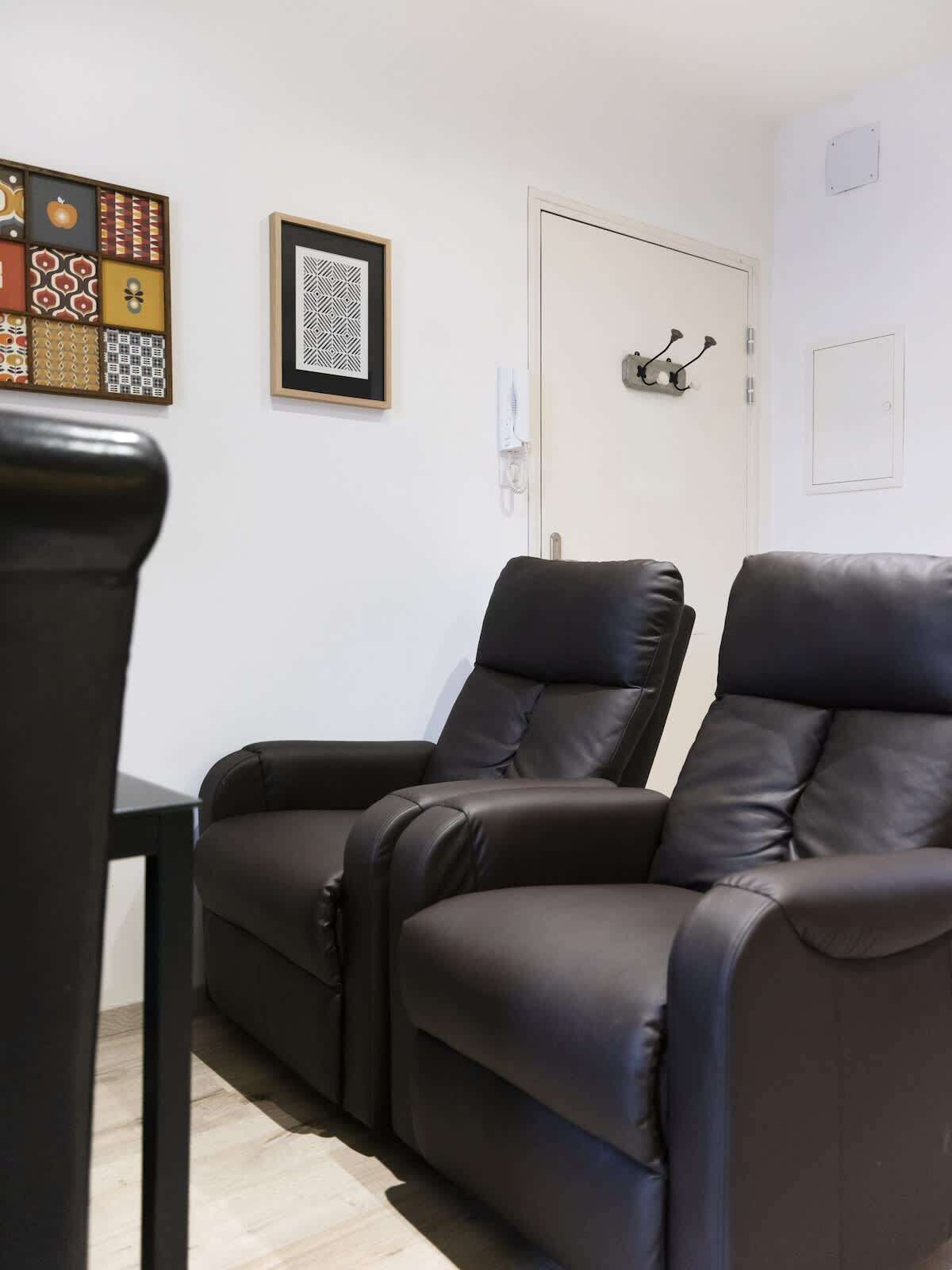 A room with two reclining chairs, a white door, a coat hook, and a framed abstract art piece on the wall.