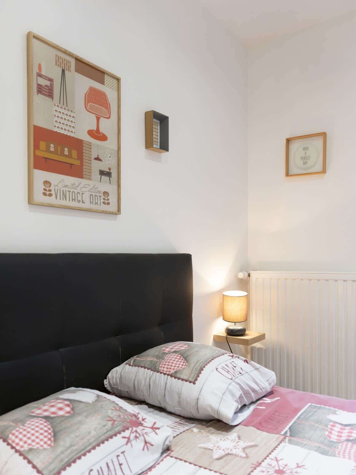 Een slaapkamer met een bed, kussens, een nachtkastje met een lamp, een lijst met een oud kunstwerk en een radiator.